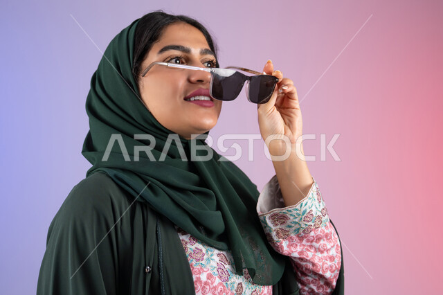 Standing up straight and confident, enjoying owning modern accessories, taking care of one's appearance and elegance, Emirati woman wearing an elegant abaya and contemporary black glasses, gestures of femininity and beauty, close-up portrait of a veiled Saudi Arabian Gulf woman trying on new sunglasses, colorful background