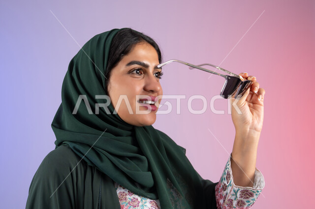 تعابير السرور والسعادة ، الاستمتاع بإمتلاك اكسسوارات عصرية, الاهتمام بالمظهر الخارجي والاناقه ،سيدة اماراتية ترتدي عباءة أنيقة ونظارة سوداء معاصرة, إيماءات الانوثة والجمال, بورترية مقرب لإمرأة عربية خليجية سعودية محجبة تقوم بتجربة النظارات الشمسية الجديدة, خلفية ملونة