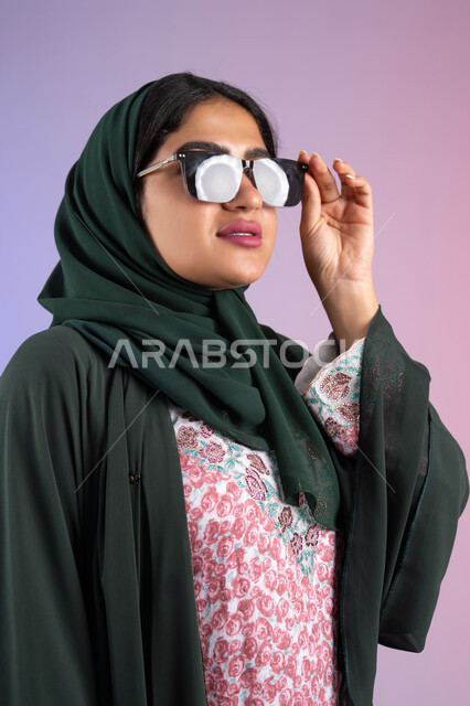 الاستمتاع بإمتلاك اكسسوارات عصرية, الاهتمام بالمظهر الخارجي والاناقه ،تعابير السرور والسعادة ، سيدة اماراتية ترتدي عباءة أنيقة ونظارة سوداء معاصرة, إيماءات الانوثة والجمال, بورترية لإمرأة عربية خليجية سعودية محجبة تقوم بتجربة النظارات الشمسية الجديدة, خلفية ملونة