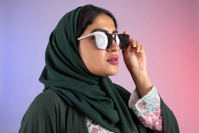 الاهتمام بالمظهر الخارجي والاناقه ، سيدة اماراتية ترتدي عباءة أنيقة ونظارة سوداء معاصرة, الاستمتاع بإمتلاك اكسسوارات عصرية, إيماءات الانوثة والجمال, بورترية مقرب لإمرأة عربية خليجية سعودية محجبة تقوم بتجربة النظارات الشمسية الجديدة, خلفية ملونة