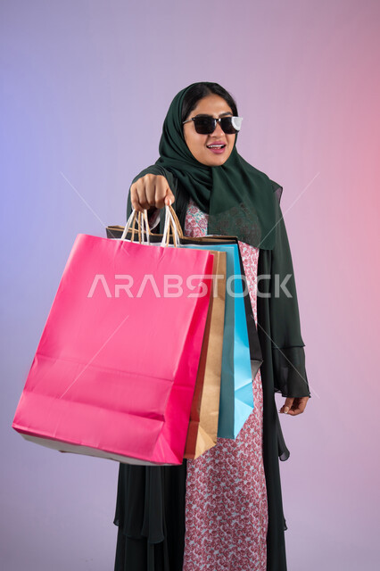 اقتناء بعض المستلزمات والهدايا، الأثر الإيجابي للتسوق على السيدات، سيدة اماراتية تستفيد من العروض الموسمية والتخفيضات، بورترية  لسيده عربية خليجية سعودية مبتسمة ترتدي العباءه والحجاب و النظارة الشمسية تحمل بيدها أكياس التسوق الملونه، خلفية ملونه