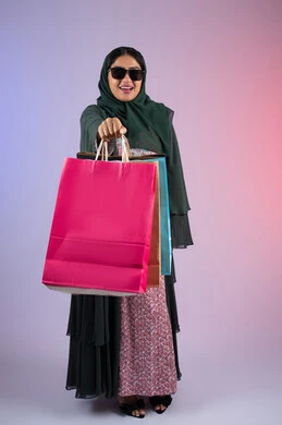 متعة التجول في أسواق ومولات الامارات العربية، الأثر الإيجابي للتسوق على السيدات، سيدة اماراتية تستفيد من العروض الموسمية والتخفيضات، بورترية  لسيده عربية خليجية سعودية مبتسمة ترتدي العباءه والحجاب و النظارة الشمسية تحمل بيدها أكياس التسوق الملونه، الجسم بالطول الكامل، خلفية ملونه