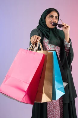 Buying necessities and clothes from shopping malls, the positive impact of shopping on women, an Emirati woman taking advantage of seasonal offers and discounts, a portrait of a smiling Saudi Arabian Gulf woman wearing an abaya and hijab, holding colorful shopping bags, a colorful background