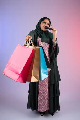 التجول وتبضع الحاجات من المراكز التجارية، ايماءات السعادة و السرور، سيدة اماراتية تستفيد من العروض الموسمية والتخفيضات، بورترية لسيده عربية خليجية سعودية مبتسمة ترتدي العباءه والحجاب تمسك بيدها النظارة الشمسية تحمل بيدها أكياس التسوق الملونه ، الجسم بالطول الكامل ، خلفية ملونه