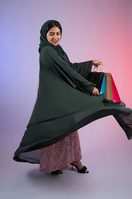 Gestures of happiness and pleasure, purchasing some supplies and gifts, an Emirati woman taking advantage of seasonal offers and discounts, a portrait of a smiling Saudi Arabian Gulf woman wearing an abaya and hijab, holding colorful shopping bags in her hand, full body, colorful background