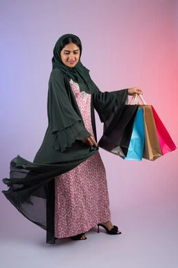 الاستمتاع في قضاء وقت ممتع في إحدى المولات التجاريه، سيدة اماراتية تستفيد من العروض الموسمية والتخفيضات، بورترية لسيده عربية خليجية سعودية مبتسمة ترتدي العباءه والحجاب تحمل بيدها أكياس التسوق الملونه ، الجسم بالطول الكامل ، خلفية ملونه