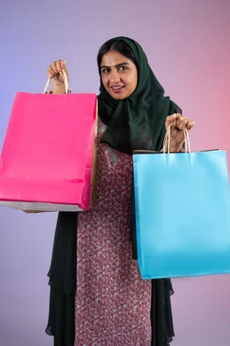 ايماءات السرور بعد التسوق والشراء وتأثيره الإيجابي على المرأة، سيدة اماراتية تستفيد من العروض الموسمية والتخفيضات، بورترية لسيده عربية خليجية سعودية مبتسمة ترتدي العباءه والحجاب تحمل بيدها أكياس التسوق الملونه ، خلفية ملونه