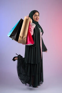 متعة التسوق في المولات التجارية،، الأثر الإيجابي للتسوق على السيدات، سيدة اماراتية تستفيد من العروض الموسمية والتخفيضات، بورترية من الجانب  لسيده عربية خليجية سعودية مبتسمة ترتدي العباءه والحجاب تحمل بيدها أكياس التسوق الملونه، الجسم بالطول الكامل ، خلفية ملونه