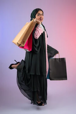 Shopping and buying supplies and gifts, the positive impact of shopping on women, an Emirati woman taking advantage of seasonal offers and discounts, a side portrait of a smiling Saudi Arabian Gulf woman wearing an abaya and hijab holding colorful shopping bags, full body, colorful background