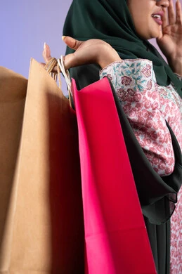 شراء المستلزمات والتبضع من المولات التجارية، سيدة اماراتية تستفيد من العروض الموسمية والتخفيضات، بورترية لسيده عربية خليجية سعودية مبتسمة ترتدي العباءه والحجاب تحمل بيدها أكياس التسوق الملونه، الأثر الإيجابي للتسوق على السيدات، خلفية ملونه