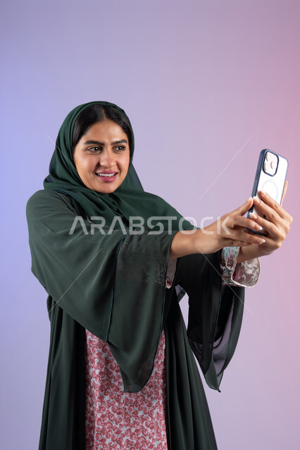 استخدام جهاز تقني حديث ومتطور، برامج وتطبيقات الجوال المتنوعة، شابة سعودية توثق اللحظات السعيدة، بورترية لشابة عربية خليجية اماراتية تمسك بيدها جهاز الهاتف المحمول تقوم بالتقاط صور السيلفي التذكارية ، الجسم بالطول الكامل، خلفية ملونه