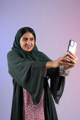 استخدام جهاز تقني حديث ومتطور، برامج وتطبيقات الجوال المتنوعة، شابة سعودية توثق اللحظات السعيدة، بورترية لشابة عربية خليجية اماراتية تمسك بيدها جهاز الهاتف المحمول تقوم بالتقاط صور السيلفي التذكارية ، الجسم بالطول الكامل، خلفية ملونه