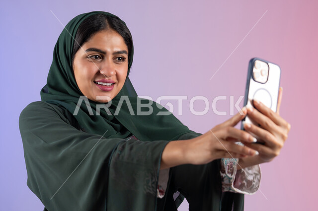 برامج وتطبيقات الجوال المتنوعة، استخدام الأجهزة التقنية الحديثة، شابة سعودية توثق اللحظات السعيدة، بورتريه مقرب ليد  شابة عربية خليجية اماراتية تمسك بيدها جهاز الهاتف المحمول تقوم بالتقاط صور السيلفي التذكارية، ، خلفية ملونه