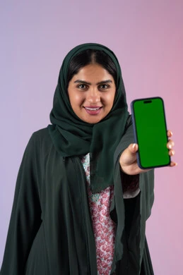 Integrating technology into practical life, displaying a blank green chroma key screen on a mobile phone, getting work done quickly and easily, portrait of an Arab Gulf Emirati woman holding a mobile phone in her hand, a Saudi woman looking at the camera with gestures of happiness and pleasure, colorful background