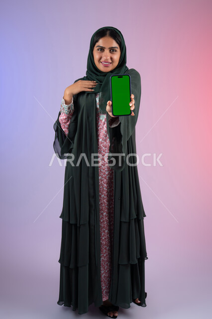 عرض شاشة كروما خضراء فارغة على الهاتف المحمول، إنجاز الأعمال بسرعة وسهولة، دمج التقنية والتكنولوجيا في الحياة العملية، بورترية لامراة عربيه خليجيه اماراتية  تحمل بيدها جوال محمول، سيده سعودية تنظر الي الكاميرا بايماءات السعادة و السرور ، الجسم بالطول الكامل، خلفية ملونه