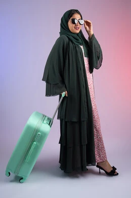 التجهيز للسفر و الرحلات الممتعة، الشعور بالسعادة والفرح, شابة سعودية تستعد لقضاء رحلة صيفية ممتعة, بورتريه لشابة عربية خليجية عمانية محجبة ترتدي العباءة الملونه و النظارة الشمسية  تمسك حقيبة السفر بإيماءات الحماس, صورة الجسم بالطول الكامل, خلفية ملونه