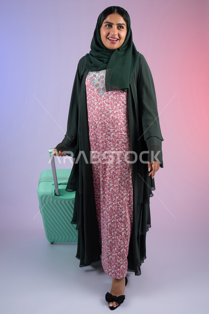 The concept of tourism and recreational tours during vacation time, feeling happy and joyful, a young Saudi woman preparing to spend a pleasant summer trip, a portrait of a veiled young Arab Gulf Omani woman wearing an abaya holding a suitcase with enthusiastic gestures, a full-length body image, a colored background