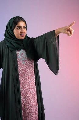 Signs that identify this place (here), identifying a specific location with cheerful gestures, portrait of a veiled young Saudi Arabian Gulf woman wearing a black abaya, an Emirati woman raising her index fingers and pointing at something, colored background