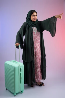 الاستعداد لقضاء رحلة صيفية ترفيهية ممتعة، الشعور بالسعادة والفرح, السياحة في إجازة الصيف, بورتريه لشابة عربية خليجية سعودية محجبة ترتدي عباءة سوداء  تمسك بيديها حقيبة السفر، امرأة اماراتية تنظر إلى الكاميرا بإيماءات الحماس و تشاور الى شيء ما, الجسم بالطول الكامل، خلفية ملونه