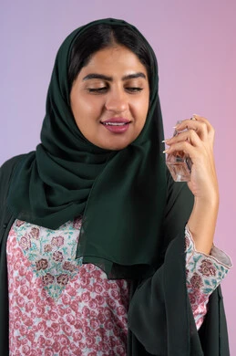 التطيب والتعطر بالعود العطري, التسويق لمنتجات العطور ، سيدة اماراتية تستخدم المستحضرات النسائية, بورترية لامرأة عربيه سعودية خليجية محجبة مبتسمة تمسك بيدها زجاجة وتقوم برش العطر، خلفية ملونة