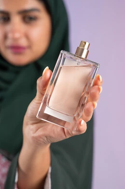 Marketing of perfume products, perfuming and fragrance with oud and aromatic oils, an Emirati woman using feminine products, a close-up portrait of a smiling veiled Saudi Arabian Gulf woman holding a bottle in her hand and spraying perfume, a colorful background