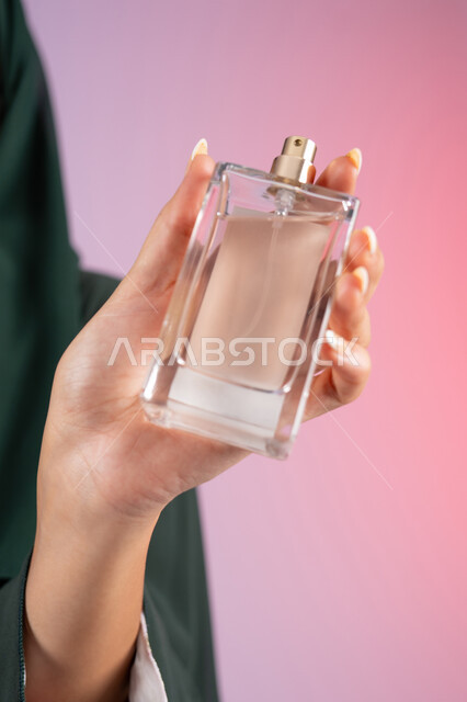 التعطر والتطيب بالعود والزيوت العطرية, التسويق لمنتجات العطور ، سيدة اماراتية تستخدم المستحضرات النسائية, بورترية مقرب لامرأة عربيه سعودية خليجية محجبة مبتسمة تمسك بيدها زجاجة وتقوم برش العطر، خلفية ملونة
