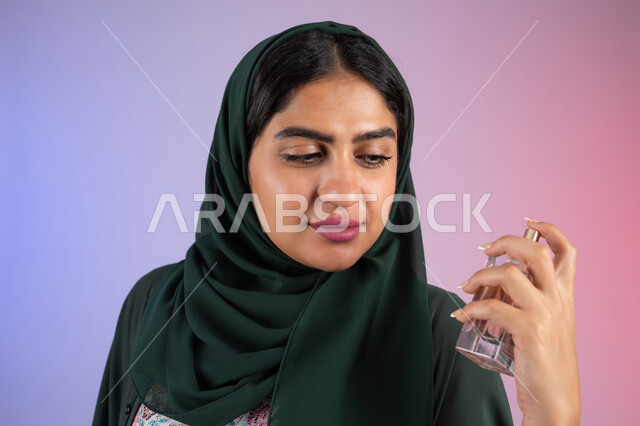 التعطر والتطيب بالعود والزيوت العطرية، سيدة اماراتية تستخدم المستحضرات النسائية, التسويق لمنتجات العطور ، بورترية مقرب لامرأة عربيه سعودية خليجية محجبة تمسك بيدها زجاجة وتقوم برش العطر، خلفية ملونة