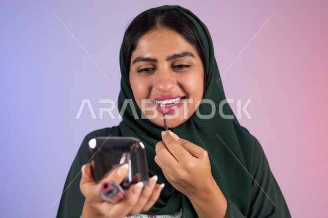 مفهوم الانوثه والاهتمام بالمظهر الخارجي، مكياج وميكاب تتوريال، سيدة اماراتية تستخدم أدوات تجميل نسائيه عصريه، التسويق لمنتجات التجميل ، بورترية مقرب لفتاه عربيه سعودية خليجيه ترتدي العباءة والحجاب تقوم بوضع احمر الشفاة ، الجسم بالطول الكامل ، خلفية ملونه