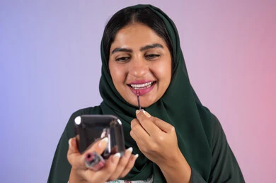 مفهوم الانوثه والاهتمام بالمظهر الخارجي، مكياج وميكاب تتوريال، سيدة اماراتية تستخدم أدوات تجميل نسائيه عصريه، التسويق لمنتجات التجميل ، بورترية مقرب لفتاه عربيه سعودية خليجيه ترتدي العباءة والحجاب تقوم بوضع احمر الشفاة ، الجسم بالطول الكامل ، خلفية ملونه