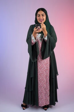 مكياج وميكاب تتوريال، سيدة اماراتية تستخدم أدوات تجميل نسائيه عصريه، مفهوم الانوثه والاهتمام بالمظهر الخارجي، التسويق لمنتجات التجميل ، بورترية لفتاه عربيه سعودية خليجيه ترتدي العباءة والحجاب تقوم بوضع احمر الشفاة ، الجسم بالطول الكامل ، خلفية ملونه