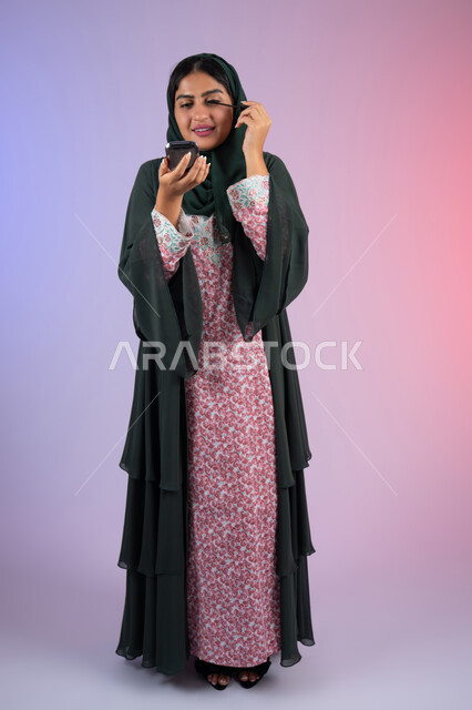 Using mascara to highlight the beauty of the eyes, taking care of beauty and outward appearance, educational courses for makeup tutorials, a Saudi expert in women's cosmetics, a portrait of an Arab Gulf Emirati woman wearing an abaya and hijab applying makeup, a full body image, a colored background