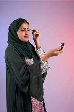 دورات تعليمية لمكياج تتوريال, الإهتمام بالجمال والمظهر الخارجي، استخدام الماسكرا لإبراز جمال العينين، خبيرة سعودية لمستحضرات التجميل النسائية، بورتريه من الجانب لامراة عربية خليجية إماراتية ترتدي العباءة والحجاب تقوم بوضع الميكاب، صورة الجسم بالكامل، خلفيه ملونه