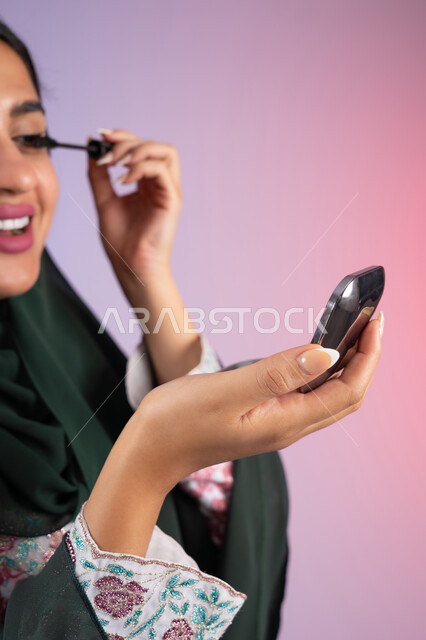 الإهتمام بالجمال والمظهر الخارجي، استخدام الماسكرا لإبراز جمال العينين، دورات تعليمية لمكياج تتوريال, خبيرة سعودية لمستحضرات التجميل النسائية، بورتريه مقرب من الجانب ليد امراة عربية خليجية إماراتية ترتدي العباءة والحجاب تقوم بوضع الميكاب، صورة الجسم بالكامل، خلفيه ملونه