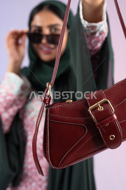 التسويق للمنتجات الاماراتية والماركات العالمية، عرض الأزياء والحقائب والاكسسوارات بالتصاميم الحديثة, متابعة صيحات الموضة، بورتريه مقرب لامراة عربية خليجية اماراتية مبتسمة ترتدي نظارات شمسية عصرية، سيده سعودية تحمل بيدها حقيبة فاخرة, خلفية ملونة