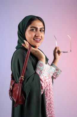The concept of fashion and elegance, modern Gulf women's clothing, a side portrait of an Arab Gulf Emirati woman wearing a colorful abaya and holding sunglasses, a Saudi woman carrying a luxurious leather bag, interest in fashion and keeping up with the latest fashion trends, gestures of self-confidence, a colorful background