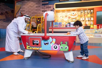 الترفيه في العطلة الصيفية، طفل خليجي يرتدي الزي الكاجوال يستمتع بلعبة الهوكي الهوائي، رجل عربي خليجي اماراتي يرتدي الكندورة و الغترة يشارك ابنه في اللعب، الأنشطة الترفيهيه الممتعة، ايماءات الاندماج والتفاعل ، قضاء الاوقات السعيدة مع العائلة، منافسة مليئة بالتحدي والإثارة