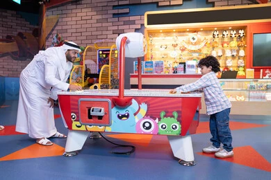 تعابير الفوز والانتصار، طفل خليجي يرتدي الزي الكاجوال يستمتع بلعبة الهوكي الهوائي، رجل عربي خليجي اماراتي يرتدي الكندورة و الغترة يشارك ابنه في اللعب، الأنشطة الترفيهيه الممتعة، ايماءات الاندماج والتفاعل ، قضاء الاوقات السعيدة مع العائلة، منافسة مليئة بالتحدي والإثارة