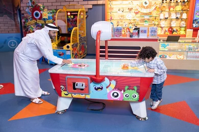 الأنشطة الترفيهيه الممتعة، ايماءات الاندماج والتفاعل ، قضاء الاوقات السعيدة مع العائلة، منافسة مليئة بالتحدي والإثارة ،رجل عربي خليجي اماراتي يرتدي الكندورة و الغترة يشارك ابنه في اللعب، طفل خليجي يرتدي الزي الكاجوال يستمتع بلعبة الهوكي الهوائي