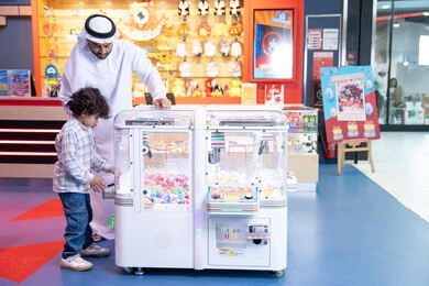 منافسة مليئة بالتحدي والإثارة ، تعابير السرور والسعادة، أجواء تفاعلية مبهجة ، انشطة صيفية ترفيهية، رجل عربي خليجي اماراتي يرتدي الكندورة و الغترة يشارك ابنه في اللعب، لحظات المرح والحماس، طفل خليجي يرتدي الزي الكاجوال يقوم  بلعب على اله المخلب