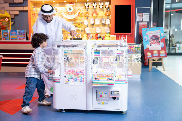 A cheerful interactive atmosphere, entertaining summer activities, gestures of integration and interaction, an Emirati Gulf Arab man wearing a kandura and ghutra participating with his son in playing, moments of fun and enthusiasm, expressions of pleasure and happiness, a Gulf child wearing casual clothes playing on the claw machine