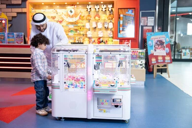 انشطة صيفية ترفيهية، ايماءات الاندماج والتفاعل، رجل عربي خليجي اماراتي يرتدي الكندورة و الغترة يشارك ابنه في اللعب، أجواء تفاعلية مبهجة ، مشاركة الأب ابنه لحظات المرح والحماس ، تعابير السرور والسعادة، طفل خليجي يرتدي الزي الكاجوال يقوم  بلعب على اله المخلب