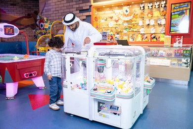 Happy summer vacation, gestures of integration and interaction, an Emirati Gulf Arab man wearing a kandura and ghutra participating with his son in playing, a cheerful interactive atmosphere, the father sharing moments of fun and enthusiasm with his son, expressions of pleasure and happiness, a Gulf child wearing casual clothes playing with the claw machine
