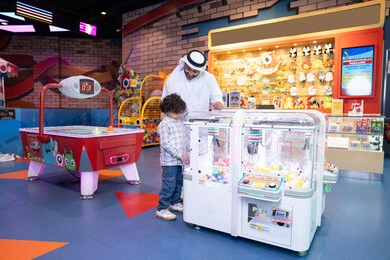 Playing recreational games, an Emirati Gulf Arab man wearing a kandura and ghutra enjoying his vacation time with his son, a cheerful interactive atmosphere, the father sharing moments of fun and excitement with his son, expressions of pleasure and happiness, a Gulf child wearing casual clothes playing with the claw machine