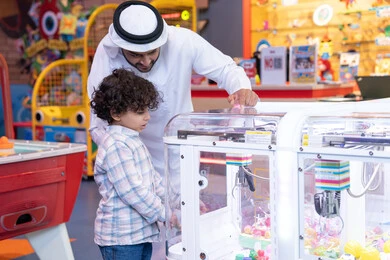 أجواء ممتعة مليئة بالمرح ، رجل عربي خليجي اماراتي يرتدي الكندورة و الغتره يستمتع بقضاء وقت العطله مع ابنه، أجواء تفاعلية مبهجة ، مشاركة الأب ابنه لحظات المرح والحماس ، تعابير السرور والسعادة، طفل خليجي يرتدي الزي الكاجوال يقوم  بلعب على اله المخلب