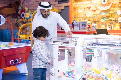 ألعاب متنوعة ومسلية، رجل عربي خليجي اماراتي يرتدي الكندورة و الغتره يستمتع بقضاء وقت العطله مع ابنه، أجواء تفاعلية مبهجة ، مشاركة الأب ابنه لحظات المرح والحماس ، تعابير السرور والسعادة، طفل خليجي يرتدي الزي الكاجوال يقوم  بلعب على اله المخلب