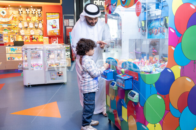 المتعة والتسلية في المراكز الترفيهية في الامارات، رجل عربي خليجي اماراتي يرتدي الكندورة و الغتره يستمتع بقضاء وقت العطله مع ابنه، أجواء تفاعلية مبهجة ، مشاركة الأب ابنه لحظات المرح والحماس ، تعابير السرور والسعادة، طفل خليجي يرتدي الزي الكاجوال يقوم  بلعب على اله المخلب