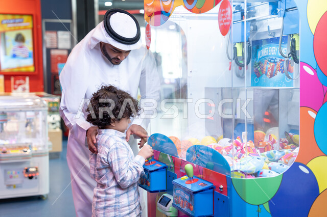الاستمتاع في اللعب في مركز الالعاب الترفيهيه، رجل عربي خليجي اماراتي يرتدي الكندورة و الغتره يستمتع بقضاء وقت العطله مع ابنه، أجواء تفاعلية مبهجة ، مشاركة الأب ابنه لحظات المرح والحماس ، تعابير السرور والسعادة، طفل خليجي يرتدي الزي الكاجوال يقوم  بلعب على اله المخلب