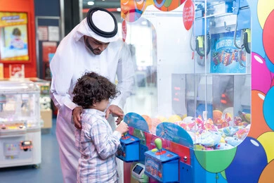 الاستمتاع في اللعب في مركز الالعاب الترفيهيه، رجل عربي خليجي اماراتي يرتدي الكندورة و الغتره يستمتع بقضاء وقت العطله مع ابنه، أجواء تفاعلية مبهجة ، مشاركة الأب ابنه لحظات المرح والحماس ، تعابير السرور والسعادة، طفل خليجي يرتدي الزي الكاجوال يقوم  بلعب على اله المخلب