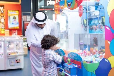 رجل عربي خليجي اماراتي يرتدي الكندورة و الغتره يستمتع بقضاء وقت ممتع مع طفله فى مدينه الملاهي، أجواء تفاعلية مبهجة ، مشاركة الأب ابنه لحظات المرح والحماس ، تعابير السرور والسعادة، طفل خليجي يرتدي الزي الكاجوال يقوم  بلعب على اله المخلب، قضاء أوقات ممتعة باللعب والتركيز
