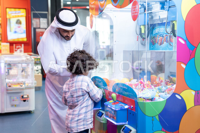 أجواء تفاعلية مبهجة ، مشاركة الأب ابنه لحظات المرح والحماس ، تعابير السرور والسعادة، طفل خليجي يرتدي الزي الكاجوال يقوم  بلعب على اله المخلب، قضاء أوقات ممتعة باللعب والتركيز، رجل عربي خليجي اماراتي يرتدي الكندورة و الغتره يستمتع بقضاء وقت ممتع مع طفله فى مدينه الملاهي